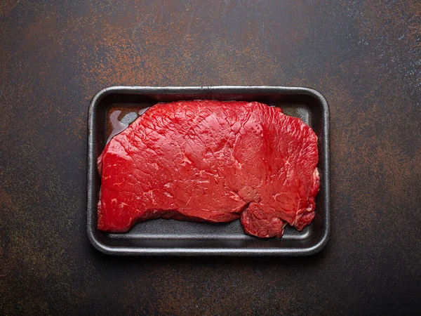 Raw uncooked top round beef steak with blood in black polystyrene tray from supermarket on dark brown rustic stone background top view, defrozing and preparing meat steak