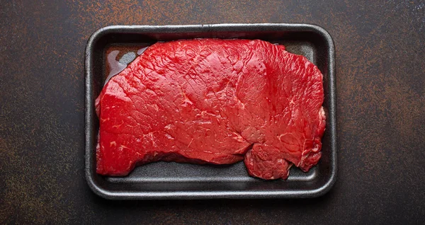 Raw uncooked top round beef steak with blood in black polystyrene tray from supermarket on dark brown rustic stone background top view, defrozing and preparing meat steak