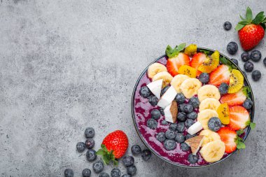 Healthy summer acai smoothie bowl with chia seeds, fresh banana, strawberry, blueberry, cocos, kiwi top view on rustic concrete background with spoon. Space for text