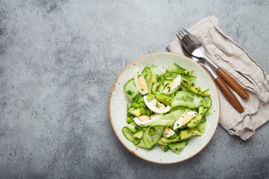 Sağlıklı yeşil avokado salata kasesi haşlanmış yumurta, dilimlenmiş salatalık, edamame fasulyesi, zeytinyağı ve seramik tabakta yeşil taşlı kırsal masa arka planında otlar. Metin için boşluk
