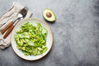 Sağlıklı vejetaryen yeşil avokado salatası kasesi dilimlenmiş salatalıklar, edamame fasulyeleri, zeytinyağı ve seramik tabakta bulunan otlar gri taş kırsal masa arka planında. Metin için boşluk