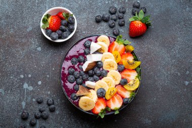 Healthy summer acai smoothie bowl with chia seeds, fresh banana, strawberry, blueberry, cocos, kiwi top view on rustic concrete background with spoon