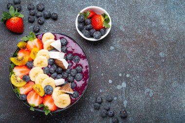 Healthy summer acai smoothie bowl with chia seeds, fresh banana, strawberry, blueberry, cocos, kiwi top view on rustic concrete background with spoon. Space for text