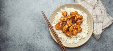 Chinese sweet and sour sticky chicken with sesame seeds and rice on ceramic plate with chopsticks top view on gray rustic stone background, traditional dish of China. Space for text