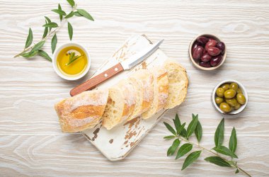 Mutfak tahtasında dilimlenmiş taze ciabatta, yeşil ve kahverengi zeytin, biberiyeli zeytin yağı, zeytin ağacı dalları, beyaz ahşap kırsal arka plan üst görünümü