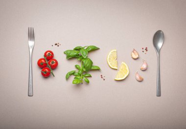 Taze kiraz domatesleri dalı, fesleğen dalı, sarımsak karanfili, limon dilimleri, mutfak kaşığı ve çatallı minimalist gri arka plan, tepe noktası.