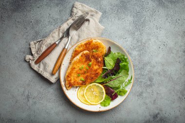 Crispy panko breaded fried chicken fillet with green salad and lemon on plate with cutlery on gray rustic concrete background table from above. Japanese style deep fried coated chicken breasts .