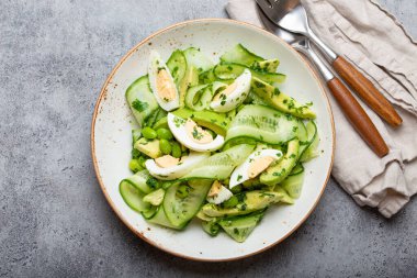 Healthy green avocado salad bowl with boiled eggs, sliced cucumbers, edamame beans, olive oil and herbs on ceramic plate top view on grey stone rustic table background