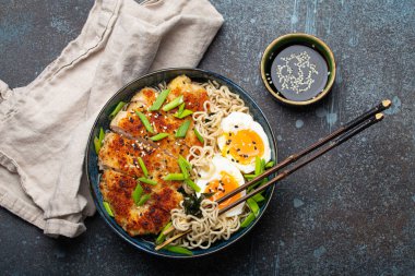 Asian noodles ramen soup with deep fried panko chicken fillet and boiled eggs in ceramic bowl with chop sticks and soy sauce on stone rustic background top view