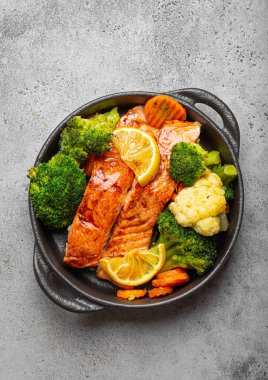 Healthy baked fish salmon steaks, broccoli, cauliflower, carrot in black cast iron casserole bowl on grey rustic stone background. Cooking a delicious low carb dinner, healthy nutrition concept