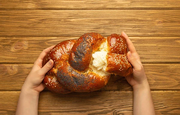 Female hands tearing off a piece of freshly baked Challah bread covered with poppy and sesame seeds, top view on rustic wooden background, traditional festive Jewish cuisine.