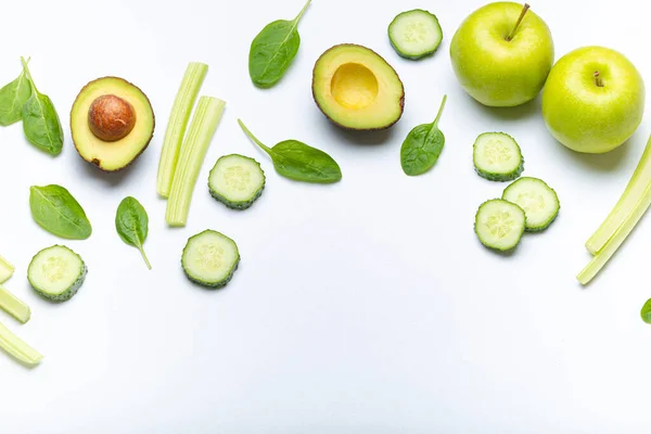 Healthy food border with green fresh vegetables and fruit on simple white background: apples, spinach, celery sticks, spinach leaves, cut avocado, cucumber. Space for text