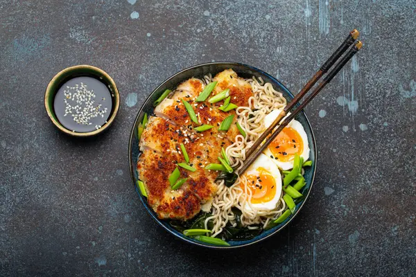 Asian noodles ramen soup with deep fried panko chicken fillet and boiled eggs in ceramic bowl with chop sticks and soy sauce on stone rustic background top view