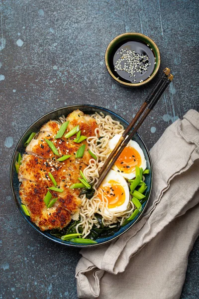 Asian noodles ramen soup with deep fried panko chicken fillet and boiled eggs in ceramic bowl with chop sticks and soy sauce on stone rustic background top view