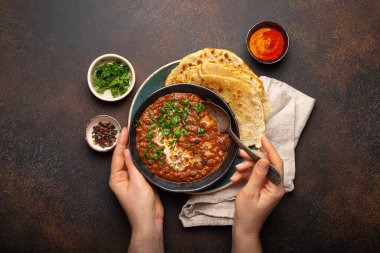 Bir kâse tutan ve geleneksel Hint Punjab yemeği yiyen kadın eller mercimek ve fasulyeli Dal Makhani naan düz ekmek ile servis edilir, kahverengi beton kırsal masa üst görüşünde taze kişniş.
