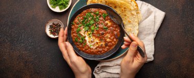 Bir kâse tutan ve geleneksel Hint Punjab yemeği yiyen kadın eller mercimek ve fasulyeli Dal Makhani naan düz ekmek ile servis edilir, kahverengi beton kırsal masa üst görüşünde taze kişniş.