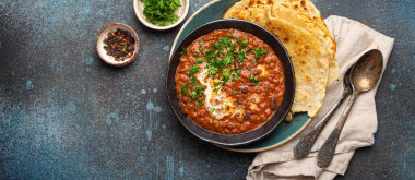 Geleneksel Hint Punjabi yemeği Dal Makhani mercimek ve siyah kasede fasulye ile birlikte naan düz ekmek, taze kişniş ve iki kaşık mavi beton kırsal masa manzaralı. Metin için boşluk.