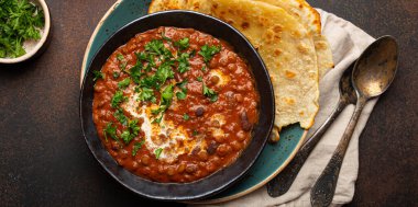 Geleneksel Hint Punjabi yemeği mercimek ve siyah kasede fasulye ile Dal Makhani naan düz ekmek, taze kişniş ve kahverengi beton kırsal masa manzaralı iki kaşık ile servis edilir..