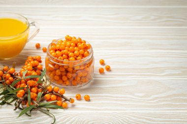 Şeffaf cam fincanda deniz dikenli sağlıklı çay, cam kavanozda olgun böğürtlen ve beyaz ahşap kırsal arka planda yapraklı dallar, deri, kalp, damar ve bağışıklık sistemi için. Boşluğu kopyala.