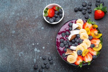 Healthy summer acai smoothie bowl with chia seeds, fresh banana, strawberry, blueberry, cocos, kiwi top view on rustic concrete background with spoon. Space for text