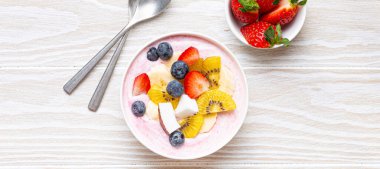 Healthy breakfast or dessert yogurt bowl with fresh banana, strawberry, blueberry, cocos, kiwi top view on rustic wooden white background with spoon