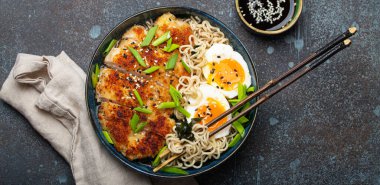 Asian noodles ramen soup with deep fried panko chicken fillet and boiled eggs in ceramic bowl with chop sticks and soy sauce on stone rustic background top view