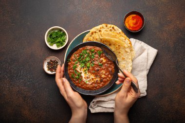 Bir kâse tutan ve geleneksel Hint Punjab yemeği yiyen kadın eller mercimek ve fasulyeli Dal Makhani naan düz ekmek ile servis edilir, kahverengi beton kırsal masa üst görüşünde taze kişniş.