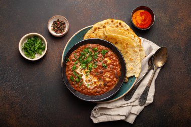Geleneksel Hint Punjabi yemeği mercimek ve siyah kasede fasulye ile Dal Makhani naan düz ekmek, taze kişniş ve kahverengi beton kırsal masa manzaralı iki kaşık ile servis edilir..