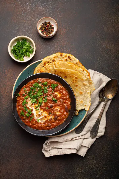 Traditional Indian Punjabi dish Dal makhani with lentils and beans in black bowl served with naan flat bread, fresh cilantro and two spoons on brown concrete rustic table top view.