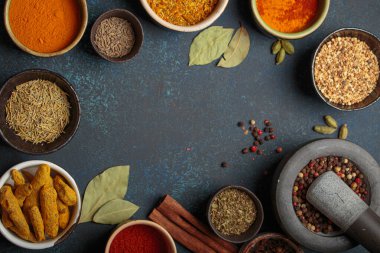 Aromatik baharat ve otlarla dolu renkli kaseler koyu bir arka planda, harç ve tokmakla bir sınır oluşturuyor. Gurme mutfağı için aromalı malzemelerin üst görünümü, kopya alanı sunar