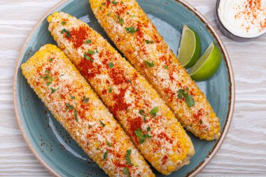 Üç ızgara mısır koçanı baharat, mayonez ve cotija peyniri ile kaplı limon dilimleri ve acı biber tozlu krema ile servis edilir.