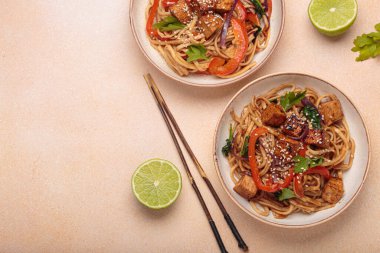 Susam tohumu ve maydanozla süslenmiş iki kase lezzetli udon eriştesi yemek çubukları ve limon dilimleriyle bej bir masada servis ediliyor.