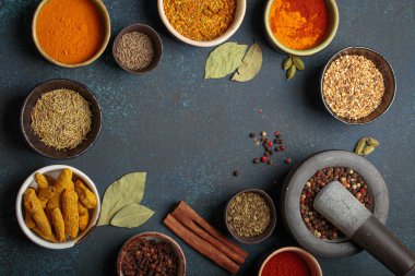 Aromatik baharat ve otlarla dolu renkli kaseler koyu bir arka planda, harç ve tokmakla bir sınır oluşturuyor. Gurme mutfağı için aromalı malzemelerin üst görünümü, kopya alanı sunar