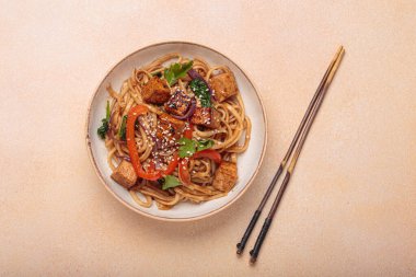 Susam tohumu ve maydanozla süslenmiş bir kase lezzetli udon eriştesi yemek çubukları ve limon dilimleriyle bej bir masada servis ediliyor.