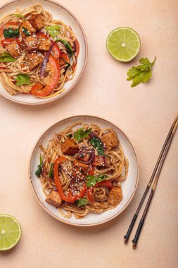 Susam tohumu ve maydanozla süslenmiş iki kase lezzetli udon eriştesi yemek çubukları ve limon dilimleriyle bej bir masada servis ediliyor.