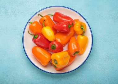 Mavi kenarlı beyaz tabakta canlı turuncu, kırmızı ve sarı dolma biberler var. Mavi arka planda bir renk patlaması var.