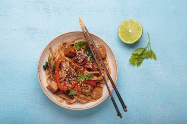 Sebzeli Udon eriştesi ve bir kasede kızarmış tofu yemek için hazır. Tofulu Asya eriştesi, ıspanak, biber, susam tohumu ve mavi ışıklı arka planda çubuklar.