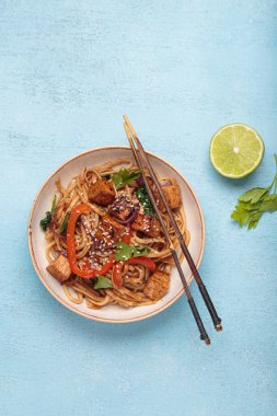 Sebzeli Udon eriştesi ve bir kasede kızarmış tofu yemek için hazır. Tofulu Asya eriştesi, ıspanak, biber, susam tohumu ve mavi ışıklı arka planda çubuklar.