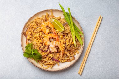 Tayland yemeği, Tayland eriştesi, karides, fıstık, limon ve yeşil soğanlı. Tabak gri bir arka planda ve yanında çubuklar var.