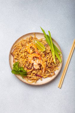 Tayland yemeği, Tayland eriştesi, karides, fıstık, limon ve yeşil soğanlı. Tabak gri bir arka planda ve yanında çubuklar var.