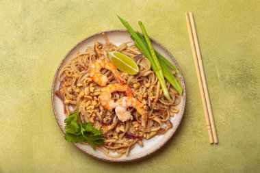 Bir kase karides, limon, fıstık ve soğanla süslenmiş Tayland eriştesi ve yanında da yeşil bir arka planda yemek çubukları. Tayland mutfağı.