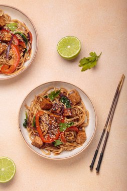 Susam tohumu ve maydanozla süslenmiş iki kase lezzetli udon eriştesi yemek çubukları ve limon dilimleriyle bej bir masada servis ediliyor.