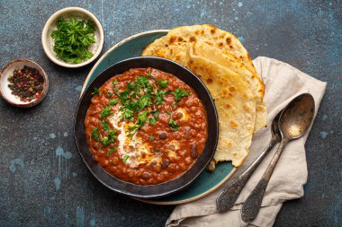 Geleneksel Hint Punjabi yemeği mercimek ve siyah kasede fasulye ile Dal Makhani naan düz ekmek, taze kişniş ve iki kaşık mavi beton kırsal masa manzaralı.