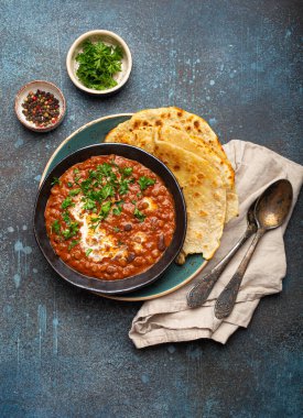 Geleneksel Hint Punjabi yemeği mercimek ve siyah kasede fasulye ile Dal Makhani naan düz ekmek, taze kişniş ve iki kaşık mavi beton kırsal masa manzaralı.