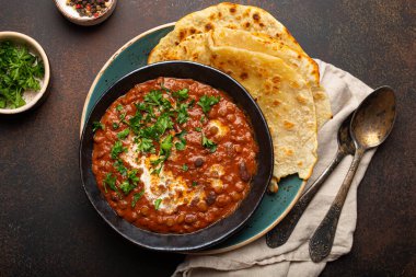 Geleneksel Hint Punjabi yemeği mercimek ve siyah kasede fasulye ile Dal Makhani naan düz ekmek, taze kişniş ve kahverengi beton kırsal masa manzaralı iki kaşık ile servis edilir..