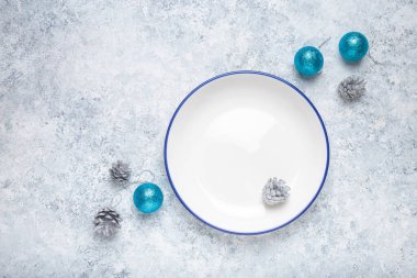 Mavi kenarlı boş beyaz tabak gümüş kozalaklar ve turkuaz noel süslemeleriyle çevrili gri desenli bir arka planda oturur ve şenlikli bir tatil masası oluşturur.