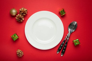 Altın kozalaklar ve küçük hediye kutuları gibi Noel süslemeleri ile çevrili kırmızı bir zemin üzerine boş beyaz tabak yerleştirilir ve bayram yemeği için şenlikli bir masa oluşturulur.
