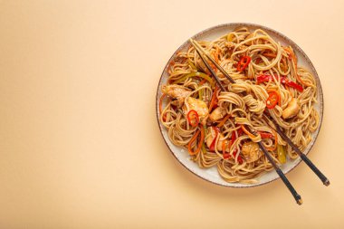 Kızarmış erişteyi teriyaki tavuğu ve sebzeyle karıştır yemek çubuklarıyla bir tabakta, bej arka plan manzarasında, boşluğu kopyala