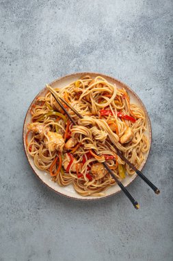 Tavuk eti, havuç, dolma biber ve diğer sebzelerle kızartılmış Udon erişteleri gri arka planda çubuklarla servis edilir ve susam tohumları serpiştirilir.