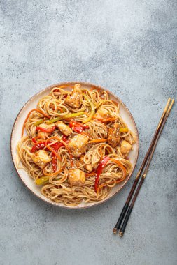 Tavuk eti, havuç, dolma biber ve diğer sebzelerle kızartılmış Udon erişteleri gri arka planda çubuklarla servis edilir ve susam tohumları serpiştirilir.